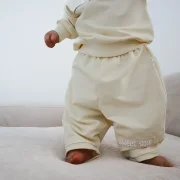 Nahaufnahme einer Baby-Beine in einer weit geschnittenen Barrel Pants mit grau-weißem Schachbrettmuster (Seersucker). Das Baby liegt auf einer hellen, strukturierten Musselindecke. Der Fokus liegt auf dem modernen Stoff und dem lockeren Schnitt am Knöchel