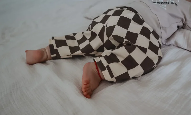 Nahaufnahme einer Baby-Beine in einer weit geschnittenen Barrel Pants mit grau-weißem Schachbrettmuster (Seersucker). Das Baby liegt auf einer hellen, strukturierten Musselindecke. Der Fokus liegt auf dem modernen Stoff und dem lockeren Schnitt am Knöchel
