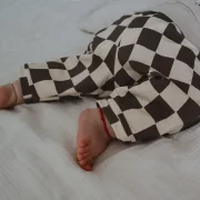 Nahaufnahme einer Baby-Beine in einer weit geschnittenen Barrel Pants mit grau-weißem Schachbrettmuster (Seersucker). Das Baby liegt auf einer hellen, strukturierten Musselindecke. Der Fokus liegt auf dem modernen Stoff und dem lockeren Schnitt am Knöchel